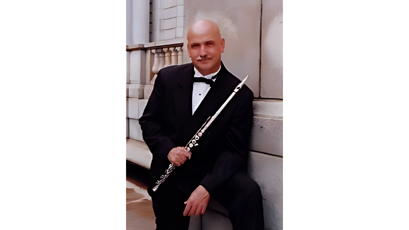Man in tuxedo holding a silver flute, leaning against a stone wall outside.