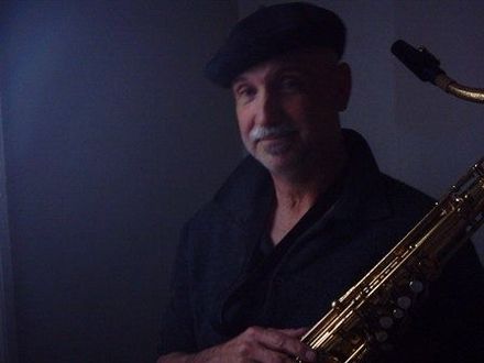 Man in tan suit holds a gold saxophone, looking up, indoors with string lights.