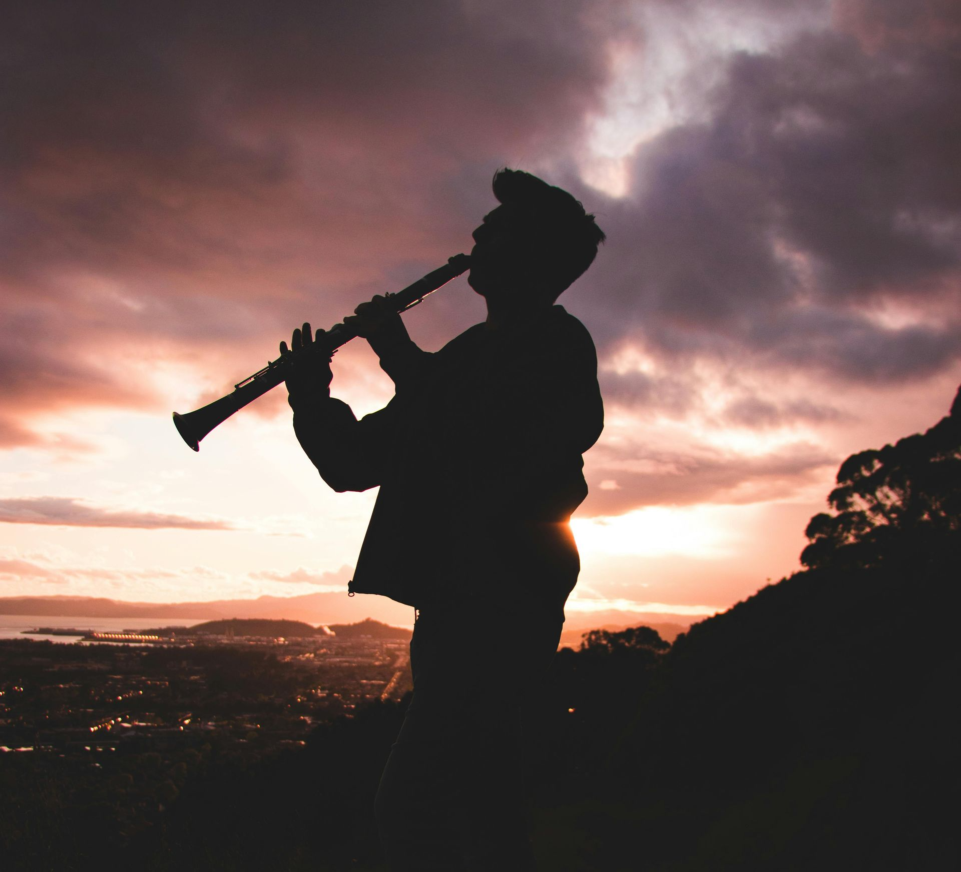 Silhouette of a person playing a clarinet against a sunset backdrop over a city.