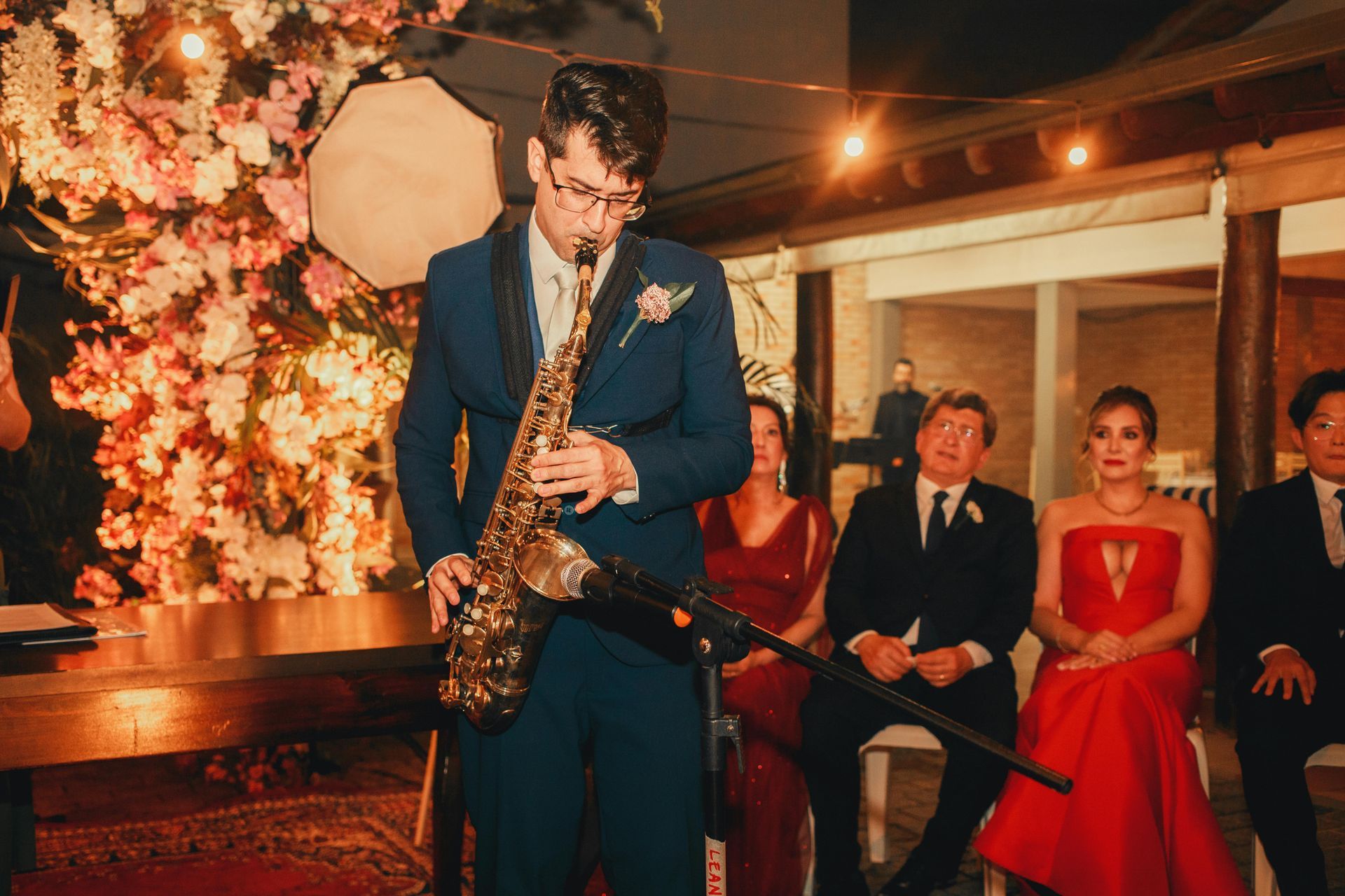 Man in blue suit plays saxophone at a wedding ceremony. Guests watch. Warm lighting, floral backdrop.