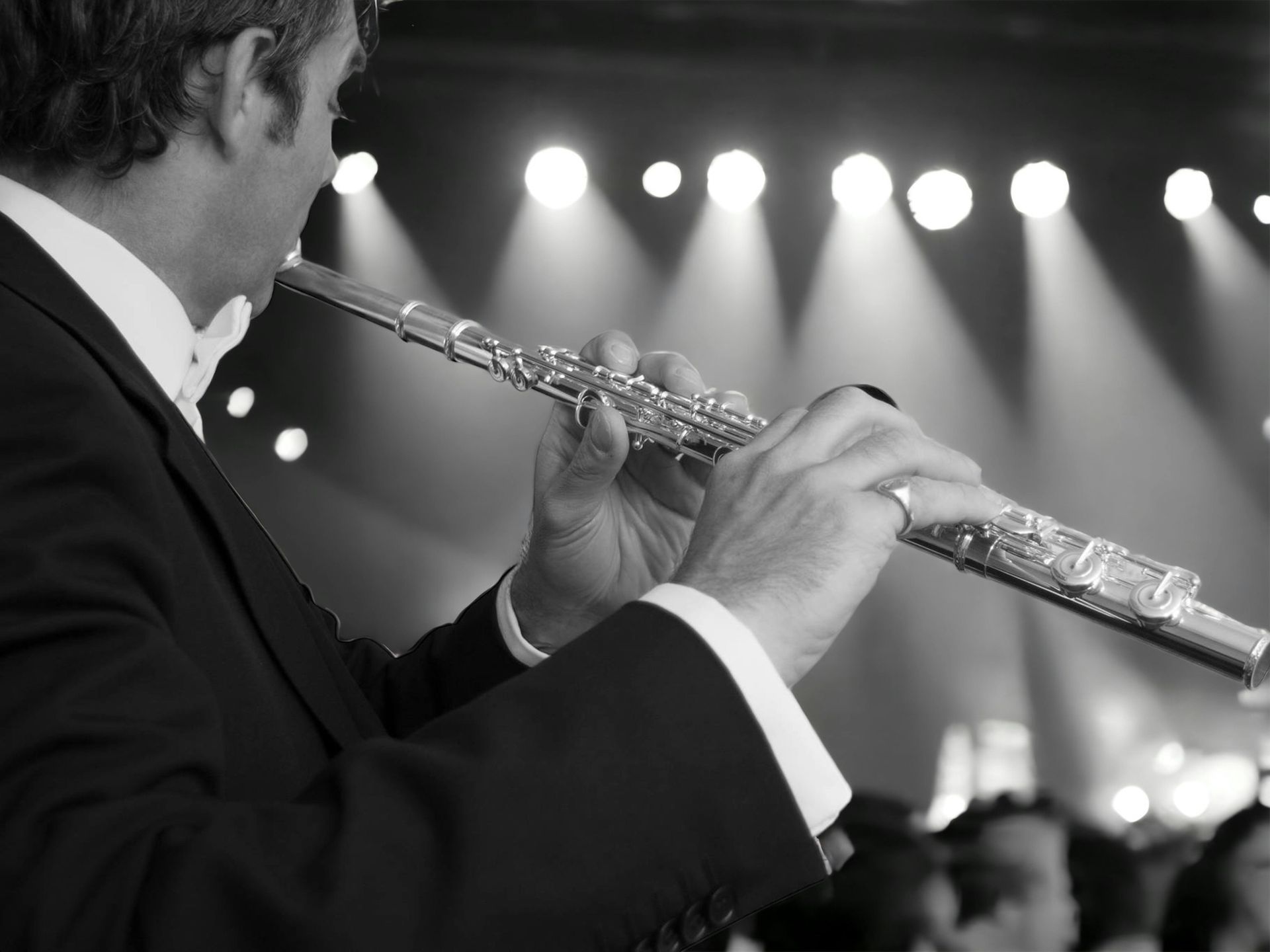 Man in tuxedo playing a silver flute onstage, lit by bright spotlights.