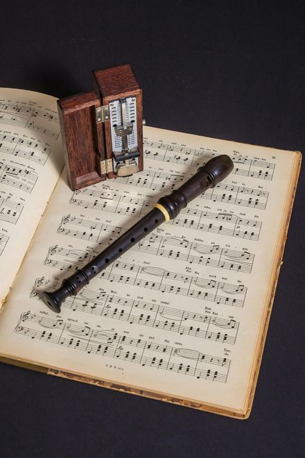 Wooden recorder and a small clock atop an open book of sheet music on a black surface.