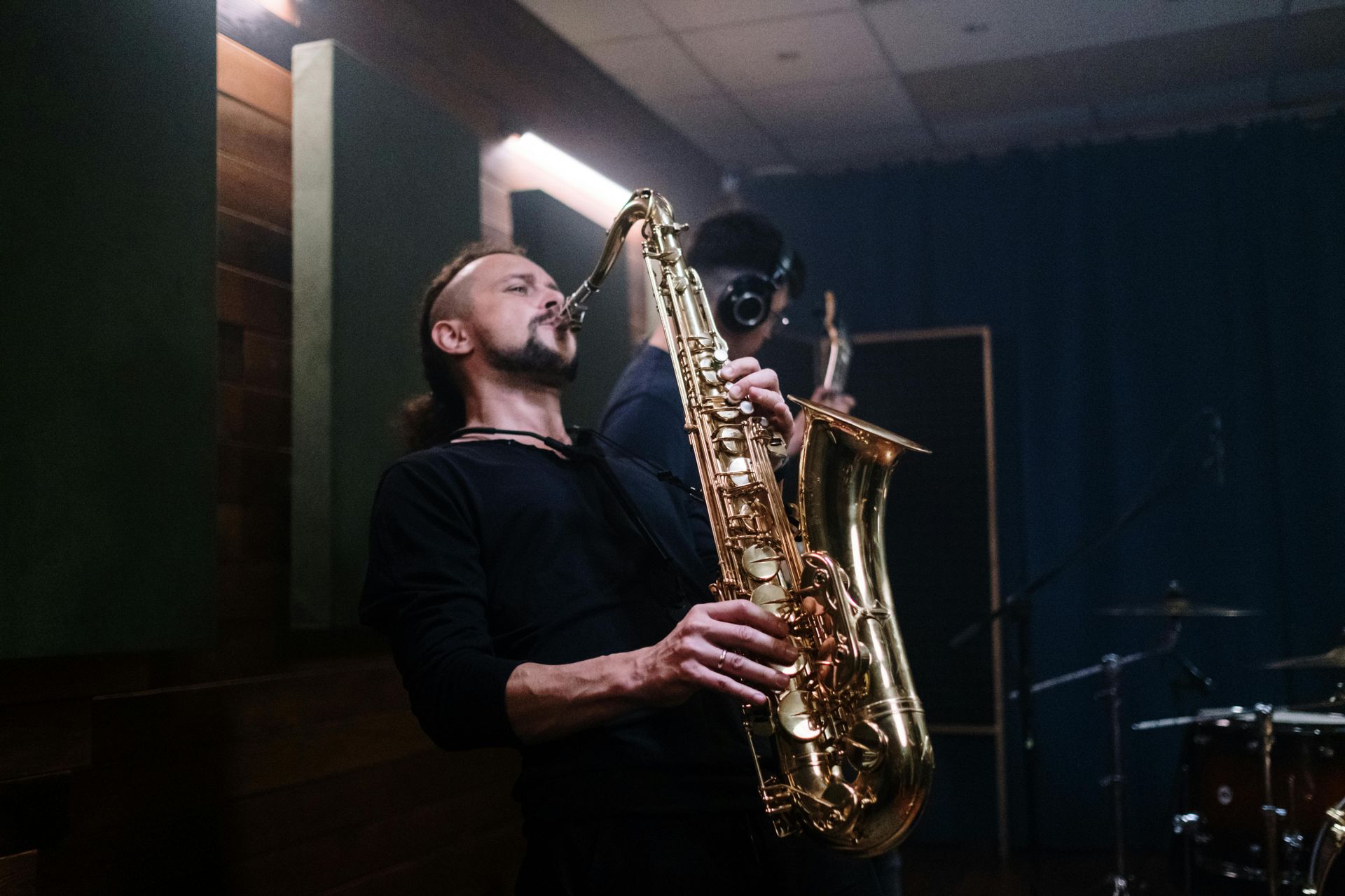 Man playing a golden saxophone in a recording studio, eyes closed, dark attire, dim lighting.