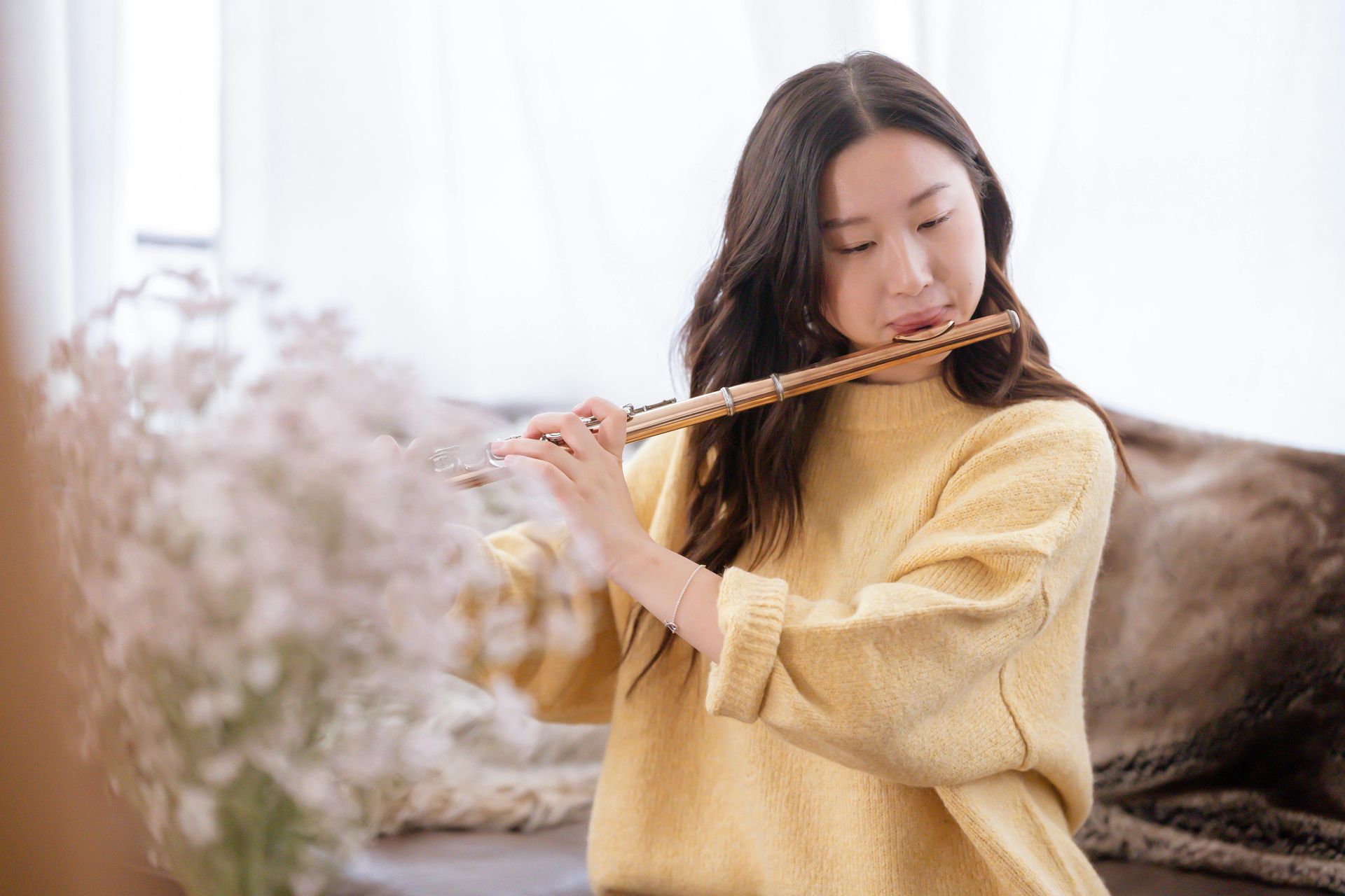 Woman in yellow sweater playing a flute indoors, with soft lighting and flowers.