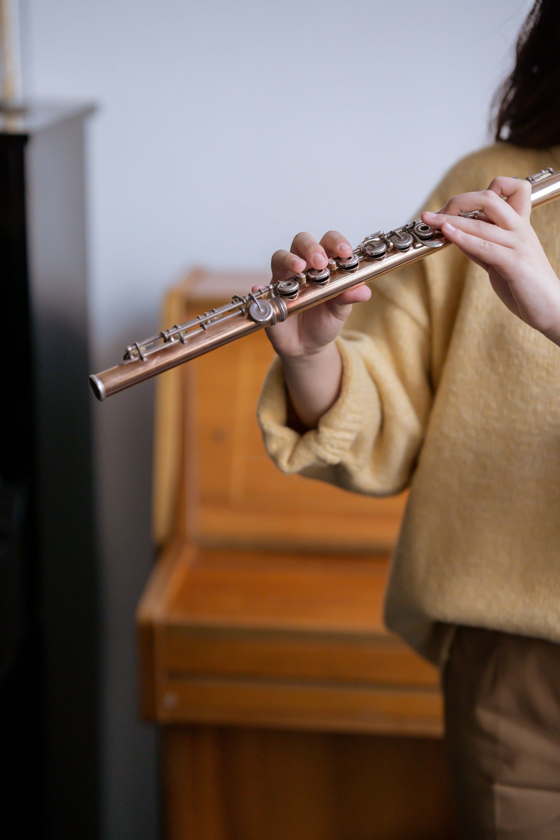 Person playing a silver flute, hands on keys. They wear a yellow sweater. Wooden piano in background.