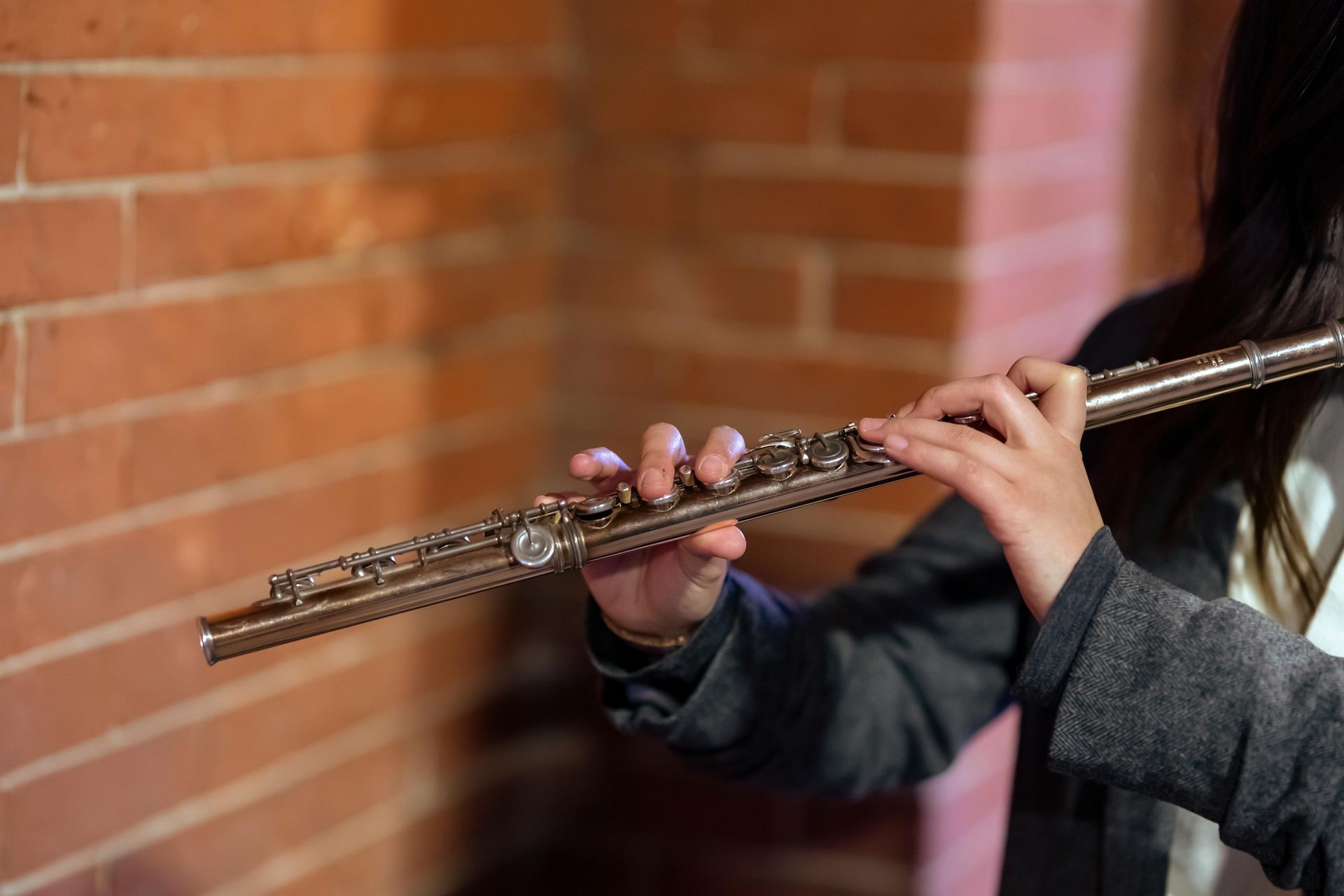 Person playing a silver flute in front of a brick wall.