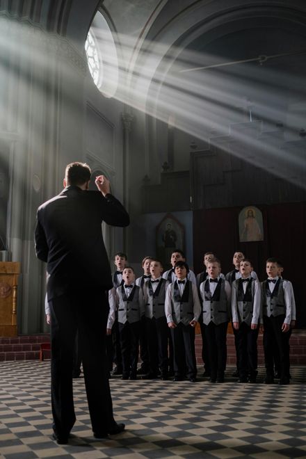 A choir conductor leading a group of young boys in a church; sunbeams shine through a window.
