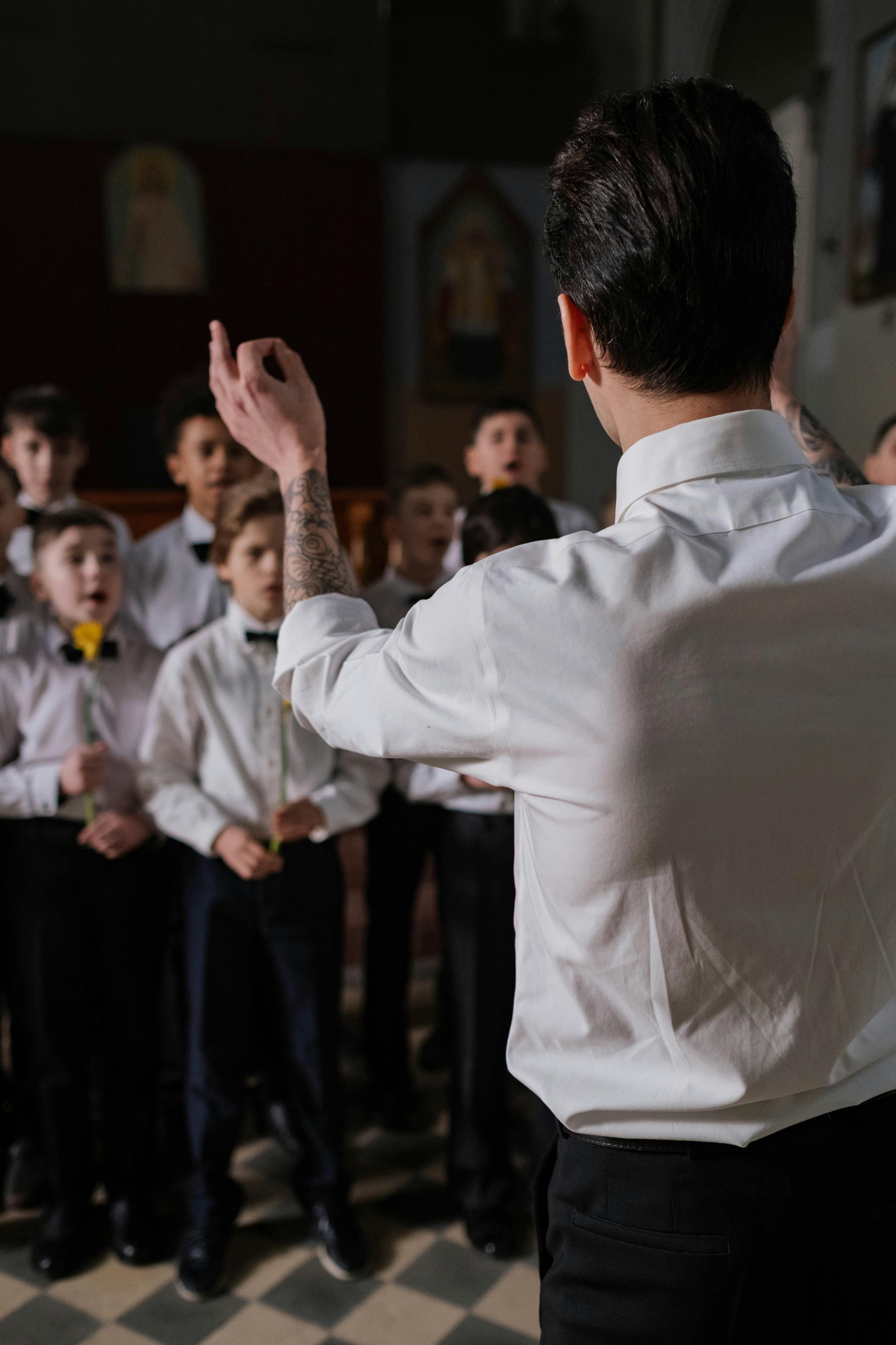 Choir conductor with tattooed arm leads boys' choir in a church. They wear white shirts and bow ties.