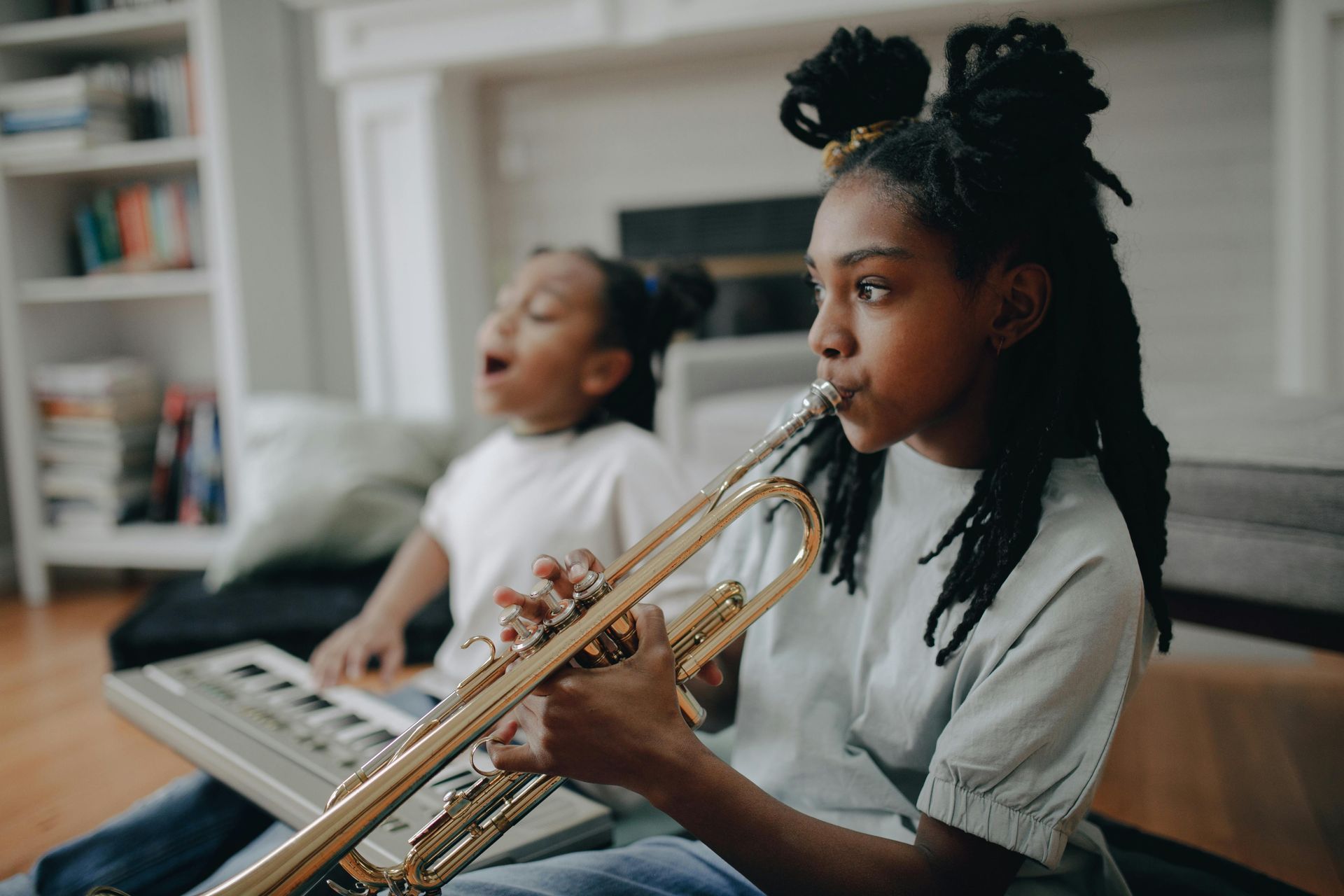 Two young Black girls playing music indoors: one on trumpet, one singing with keyboard.