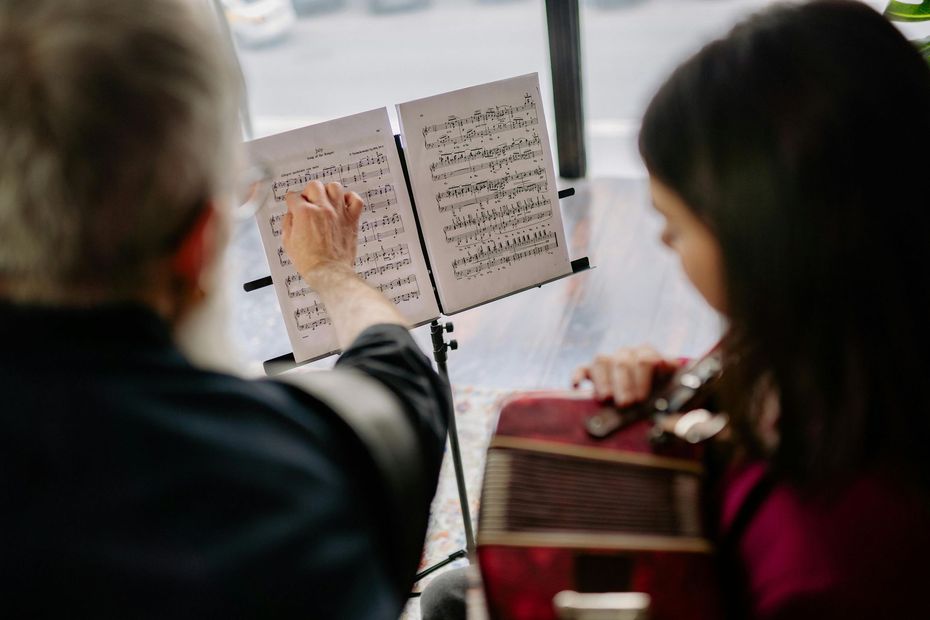 Man with gray beard points to music score for a young woman holding an instrument, indoors.