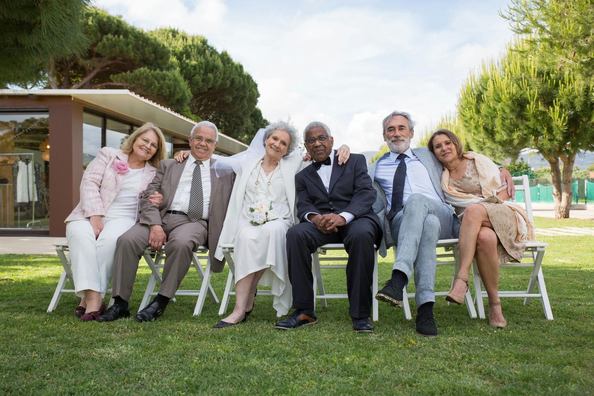Group of six people, smiling and seated on chairs on grass at an outdoor event.