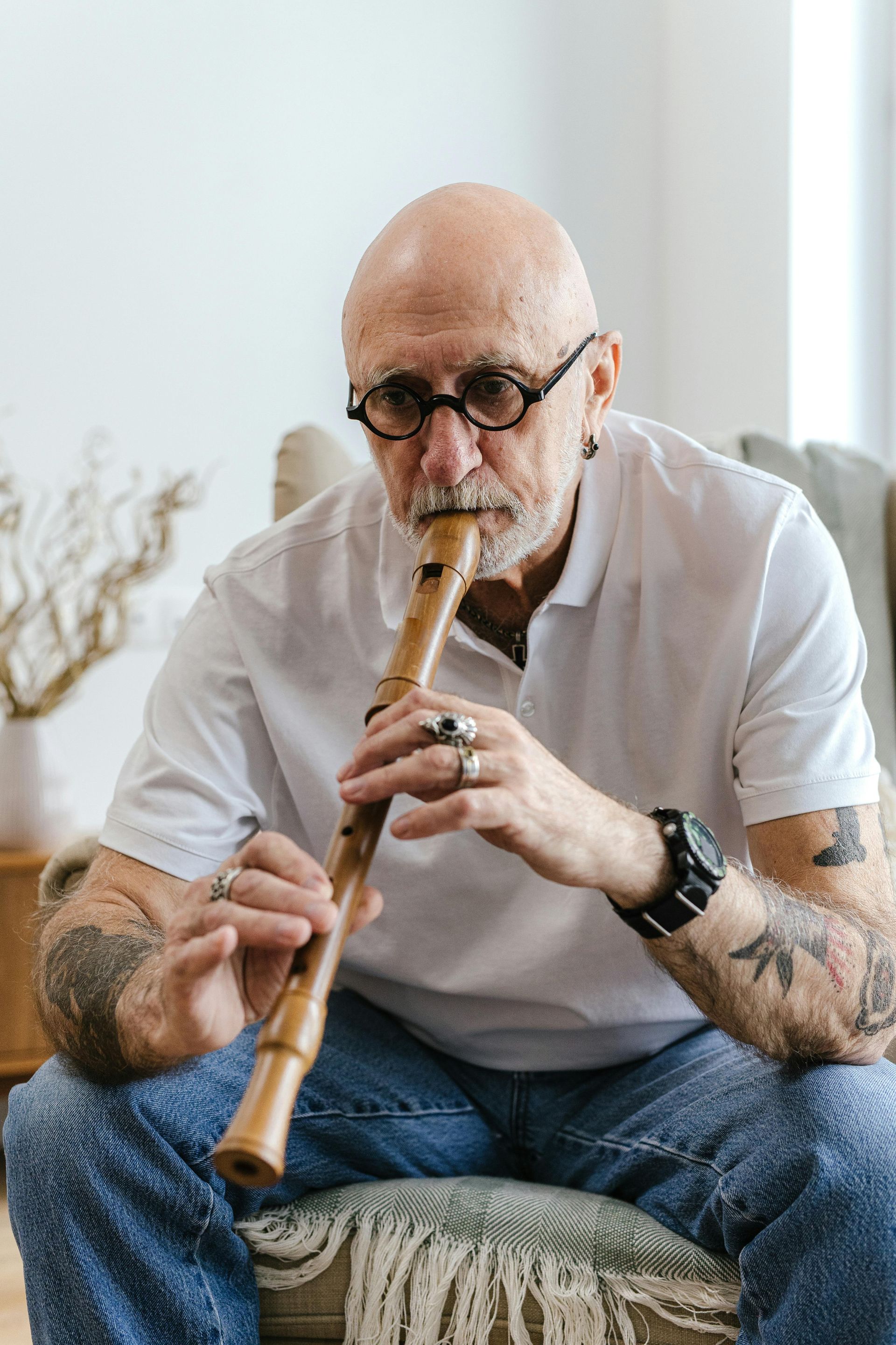 Bald man with glasses plays a wooden flute, sitting on a patterned chair; tattoos visible.