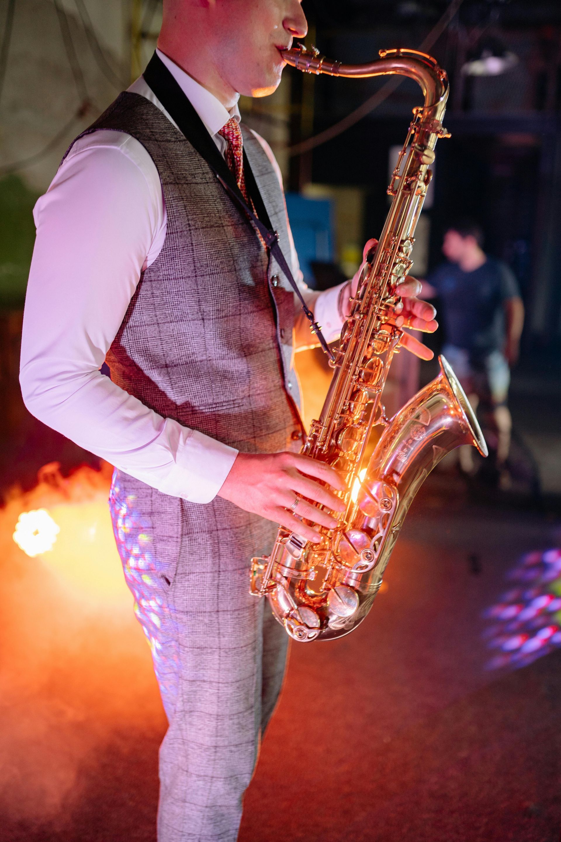 Man in vest and tie plays golden saxophone, lit by stage lights.