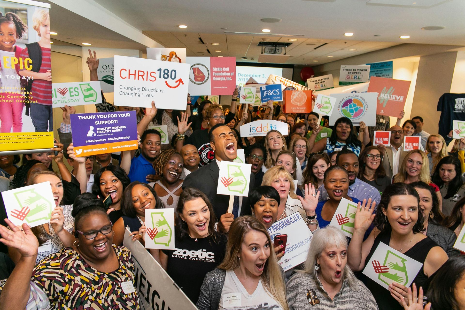 A large, diverse group of people cheering, holding signs, indoors. Many smiling, waving, with positive expressions.