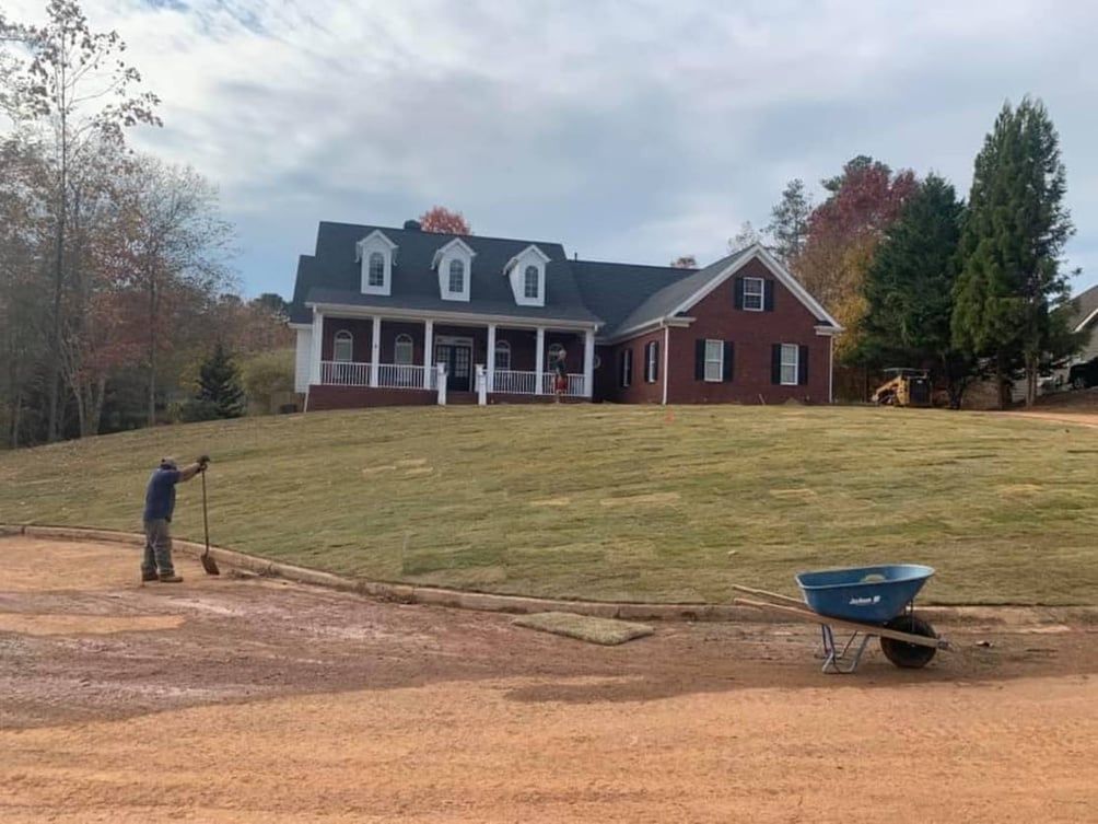 Man raking dirt, near a house with a large lawn and cloudy sky. A wheelbarrow sits nearby.