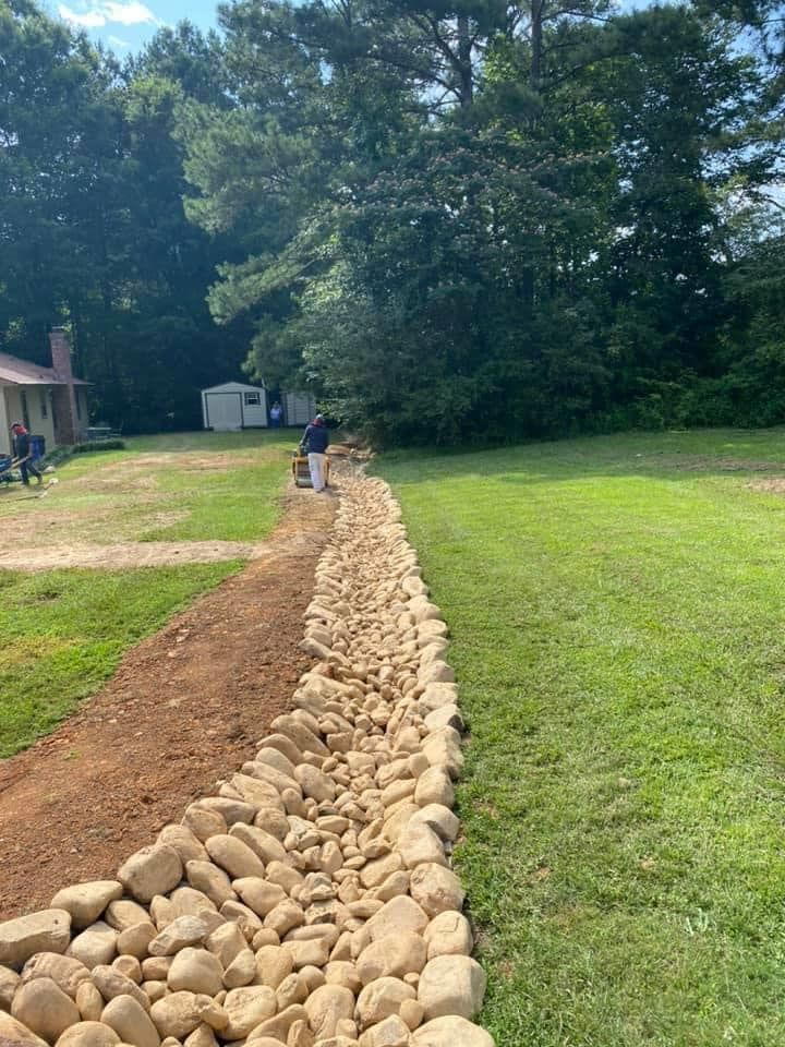 A rock-lined trench in a grassy yard, with a person working in the background near a shed and trees.