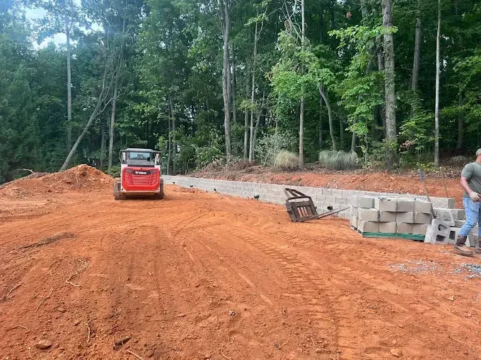 Construction site with red dirt, a Bobcat, concrete blocks, and a man. Trees in the background.