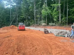 Construction site with a skid steer, blocks, and a man near a wooded area. Red dirt covers the ground.