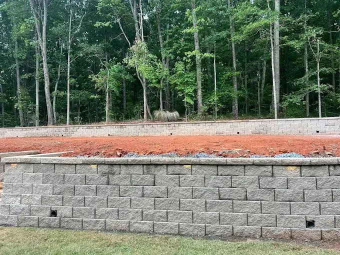 Retaining wall made of gray bricks, brown soil, and a wooded background.