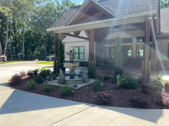 Fountain in a landscaped bed in front of a house with a covered entryway, trees and blue sky.