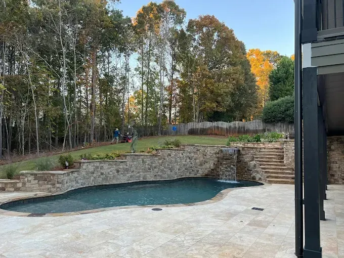 Backyard pool with stone wall, steps, and lawn, trees in background, people visible on lawn.