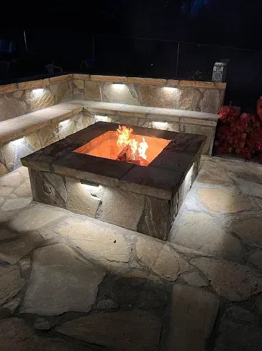 Stone fire pit with built-in seating illuminated at night, flames burning brightly.