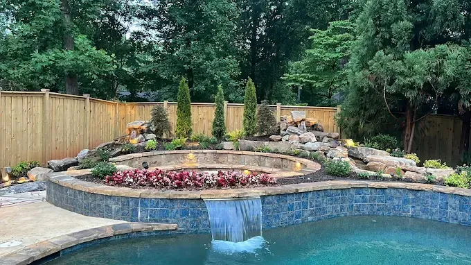 Backyard pool with a waterfall feature, fire pit, and landscaping, against a wooden fence.