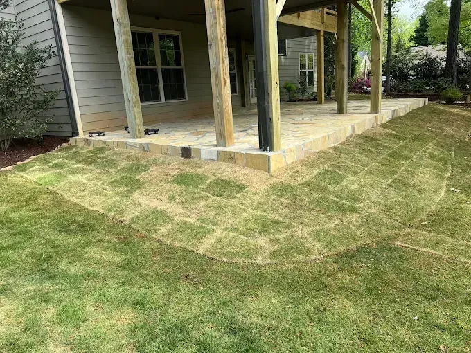 Backyard with new stone patio under a porch, surrounded by newly laid sod and existing grass.
