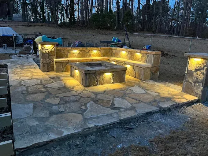 Stone patio with built-in seating around a lit fire pit. Overhead lights illuminate the space in a backyard setting.