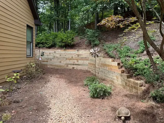 A gravel path leads to a tiered retaining wall garden. The wall is made of weathered wood blocks; trees surround.