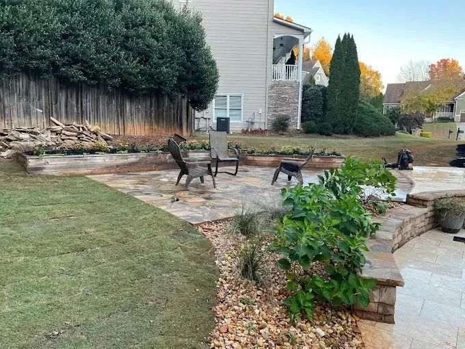 Backyard with stone patio, retaining walls, seating, and landscaping. A house in background with trees.