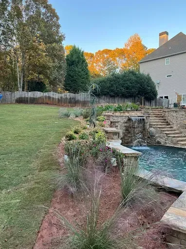 Backyard with a pool, waterfall, landscaping, and house. Green grass, blue water, and brown stone.