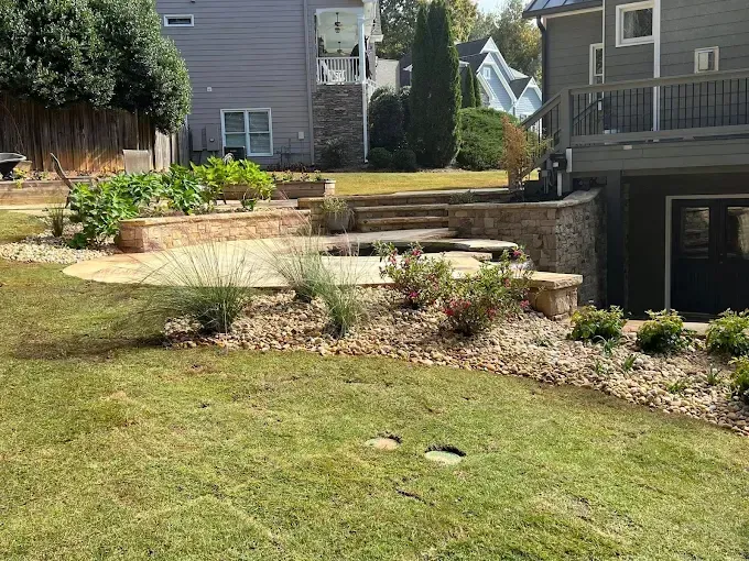 Backyard with tiered stone retaining walls, patio, and lawn. Plants and houses visible.