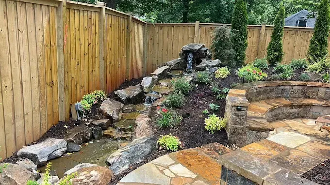 Backyard with wooden fence, stone waterfall, and patio with steps, plants.