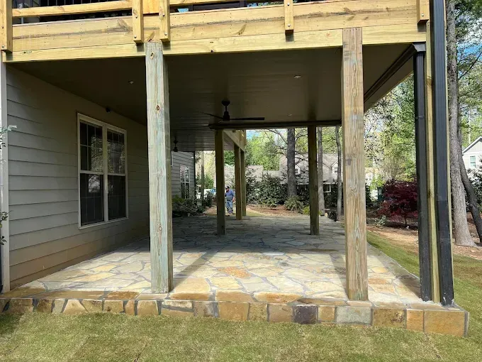 Stone patio beneath a wooden deck, supported by posts.  Exterior view with lawn and trees.