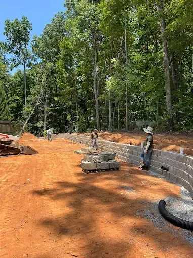 Construction workers building a retaining wall on a sunny, dirt lot surrounded by trees.