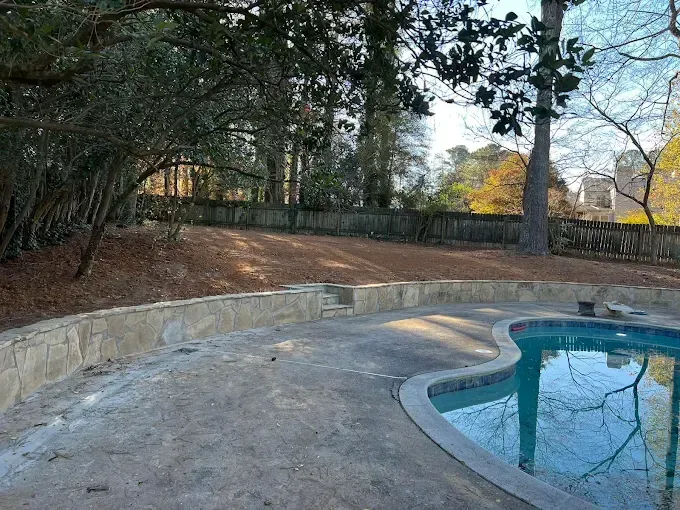 Backyard with pool, retaining wall, and trees. Concrete patio, dirt, and a wooden fence.