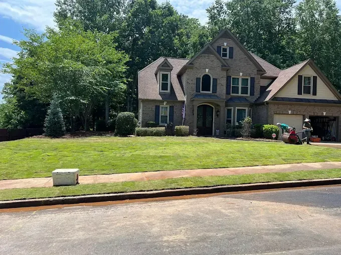 A two-story brick house with a well-maintained lawn. Trees line the background under a blue sky.