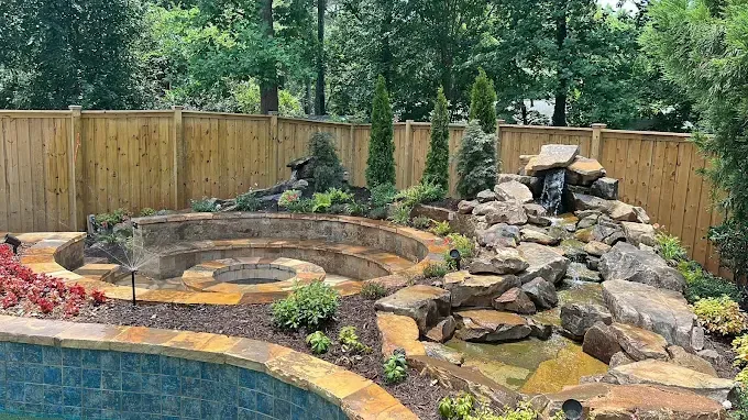 Backyard landscaping with a stone fire pit, waterfall, and pool, surrounded by a wooden fence and greenery.