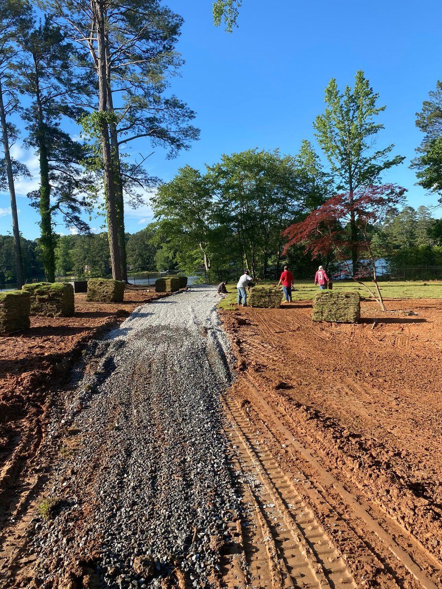 Gravel pathway construction with hay bales, surrounded by trees and workers in a sunny outdoor setting.