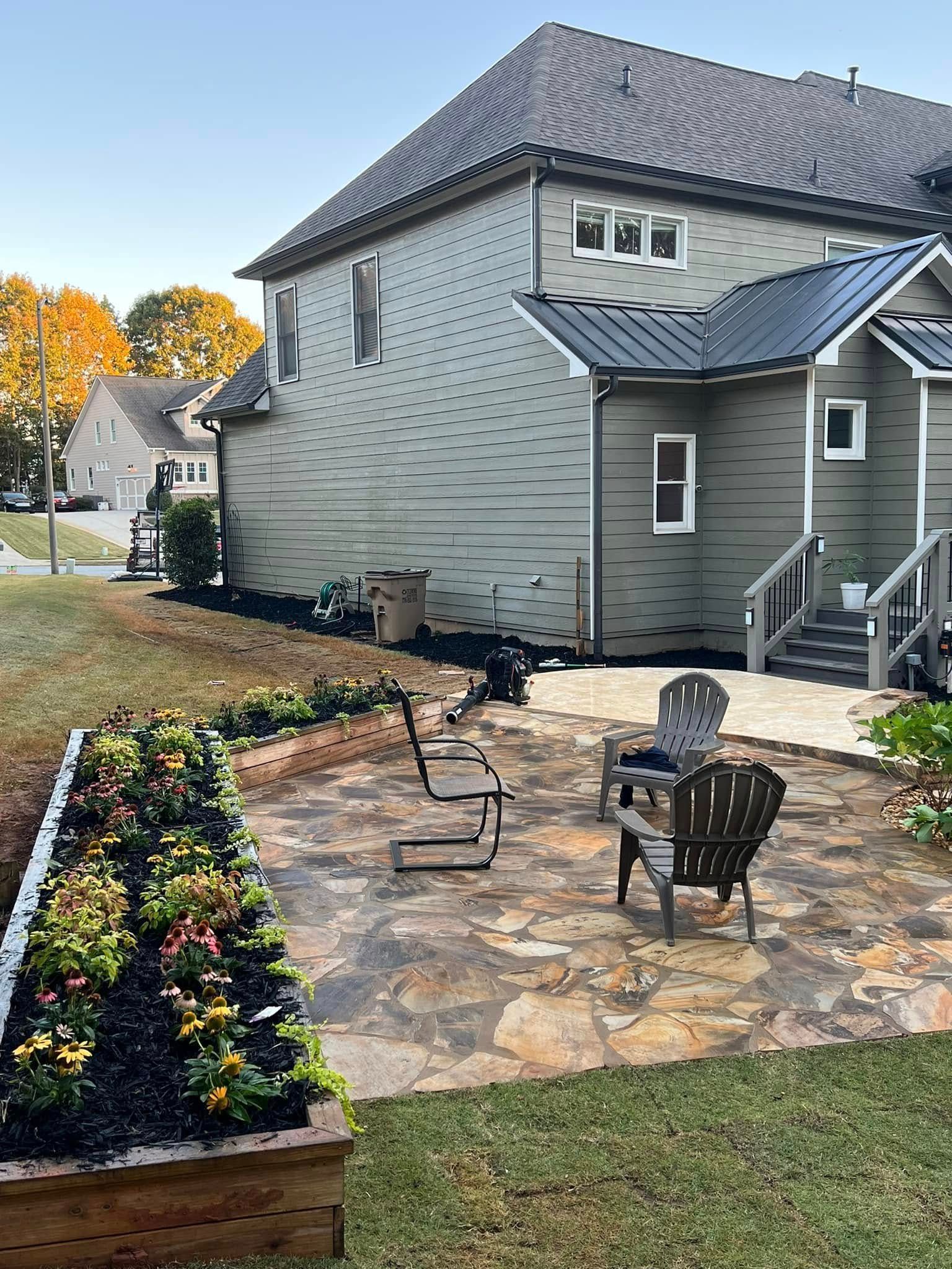 Backyard patio with chairs, a flower bed, and a two-story house with green siding.