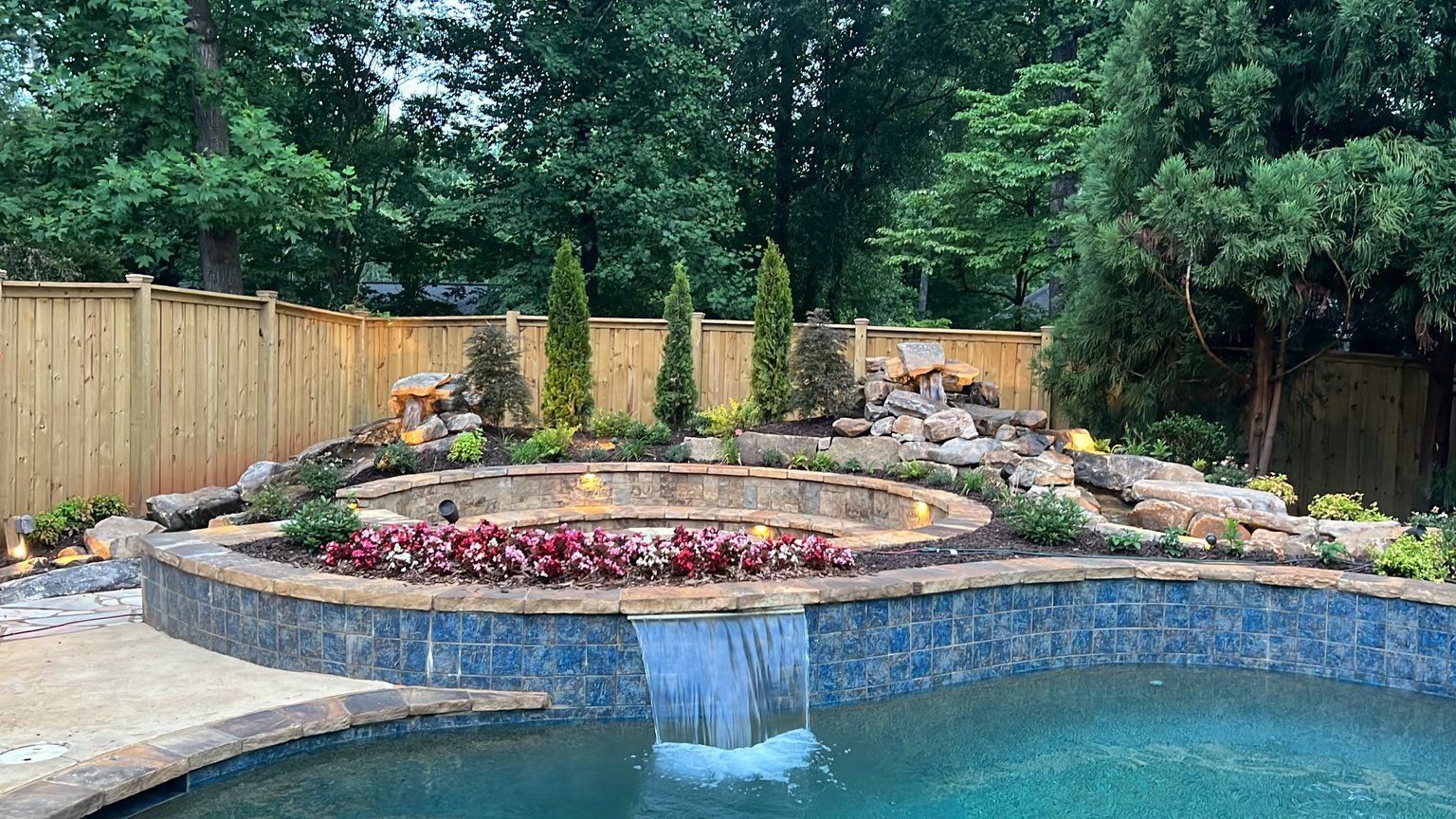 Backyard pool with waterfall, fire pit, and stone landscaping; trees and wooden fence in the background.