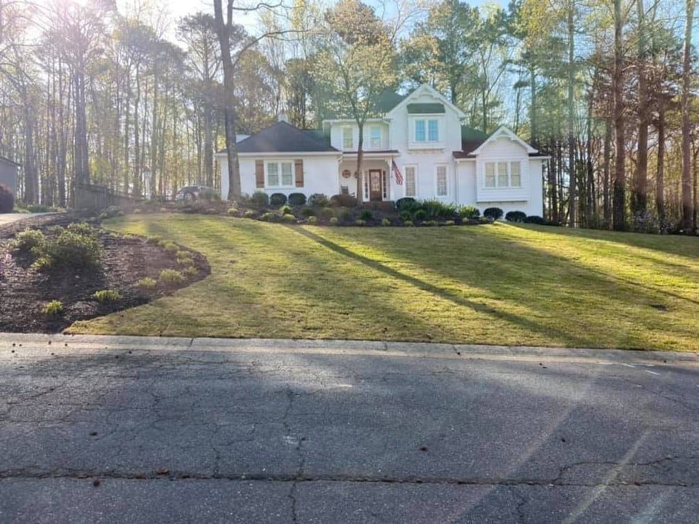White house with shutters, surrounded by green lawn and trees, viewed from a street.