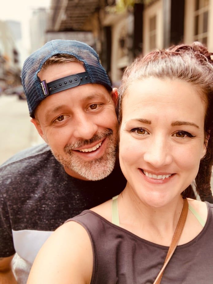 Couple smiling, posing close together outdoors. Man in trucker hat, woman in black top, smiling.