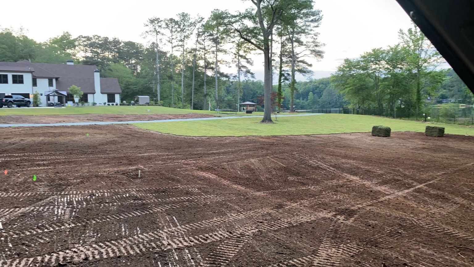 A freshly tilled brown field with tire tracks, a house in the background, and hay bales.