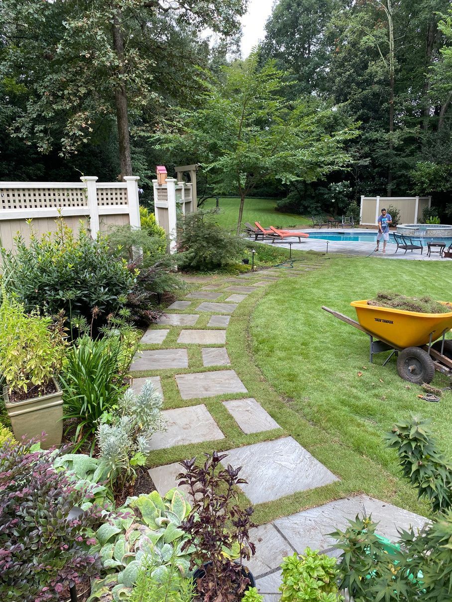 Stone path through a garden, leading to a pool area. A person relaxes poolside. Yellow wheelbarrow on the grass.