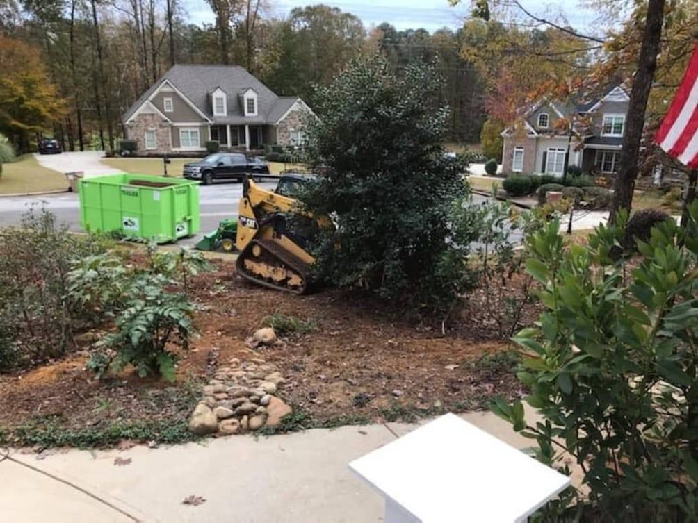 Small bulldozer removing a bush from a yard, green dumpster in the background, houses nearby.