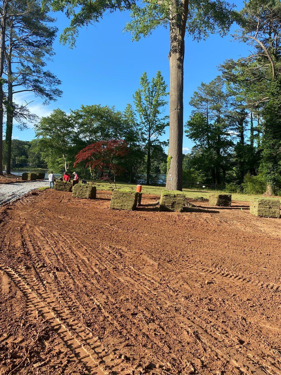 A red dirt landscape with sod laid out, trees in the background, blue sky.