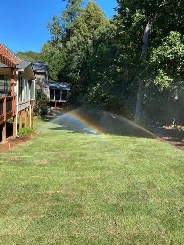 Lawn being watered with a sprinkler, creating a rainbow in a sunny yard with a house in the background.
