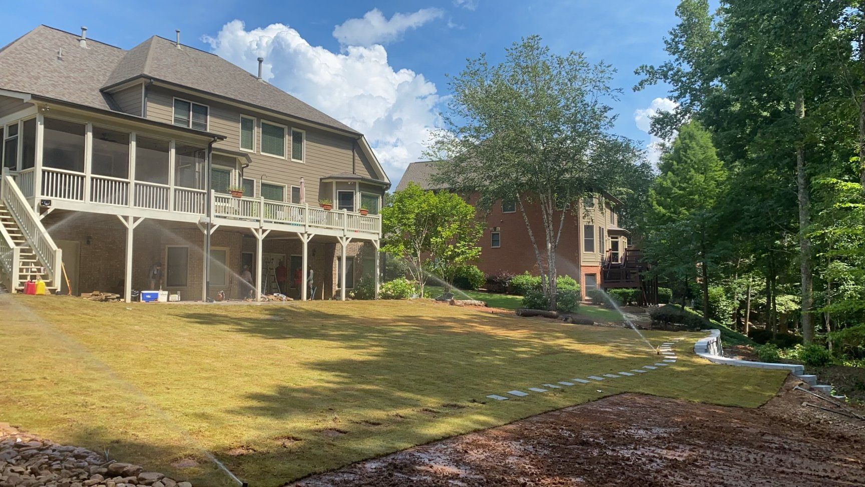 Backyard with house and trees, sprinkler system watering grass. Blue sky with some clouds.